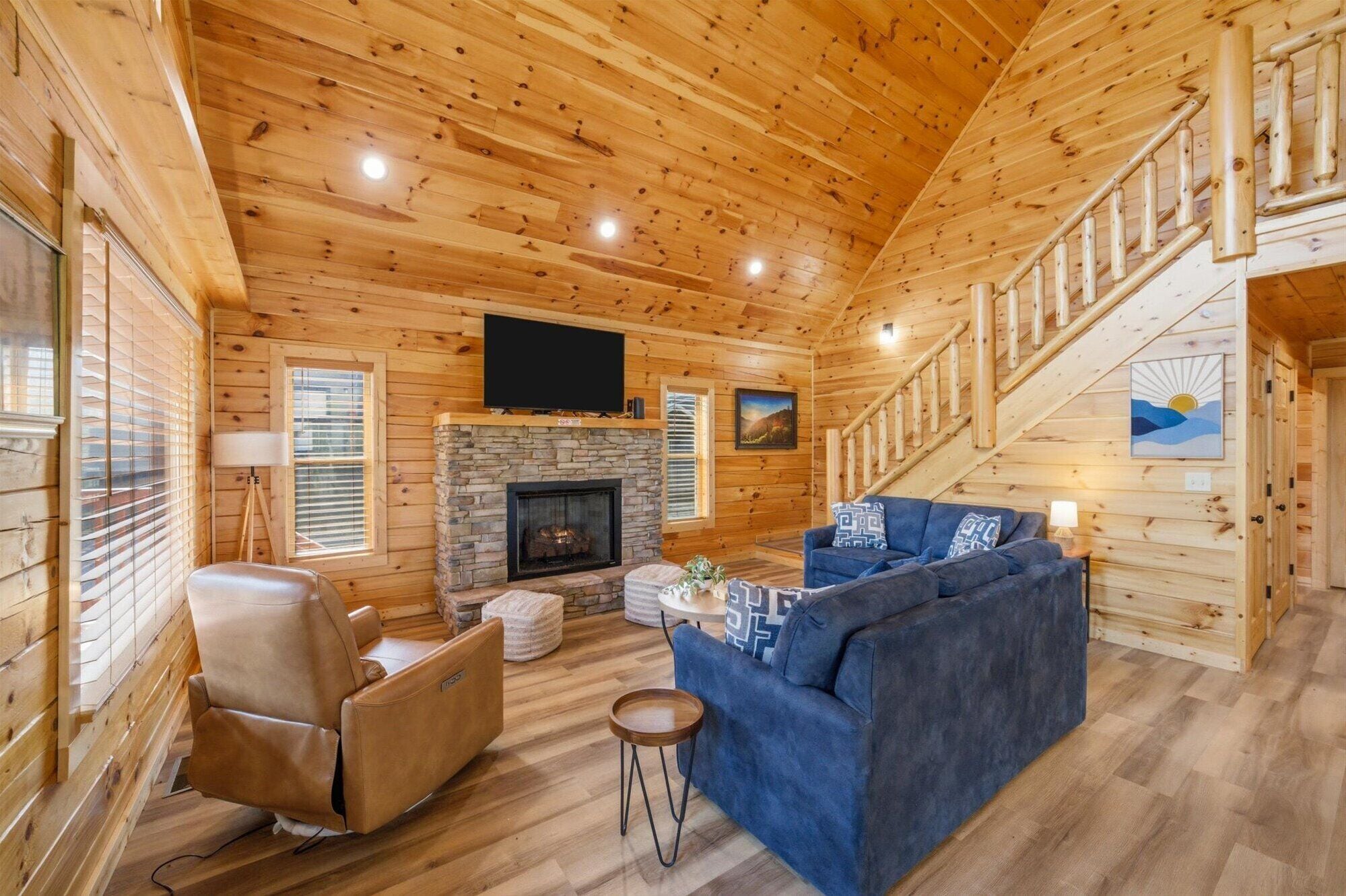 A log cabin interior with a stone fireplace, a blue sofa, a leather recliner, and a staircase.