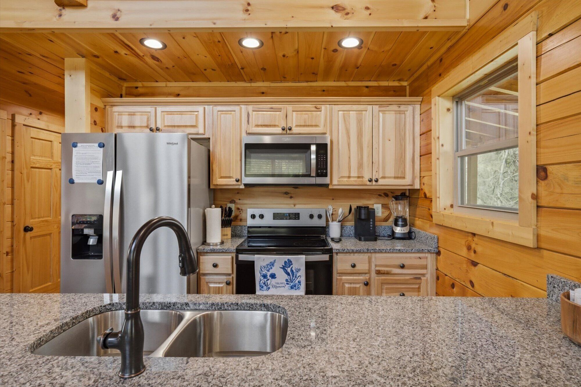 A kitchen with wooden cabinets, a stainless steel refrigerator, a microwave, an oven, a sink, granite countertops, and a window.