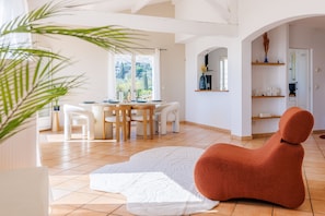 A modern interior with a dining area, a unique orange chair, and a view through a window. - La Cabanette – Villa with pool and seaview (Tourrettes-sur-Loup)