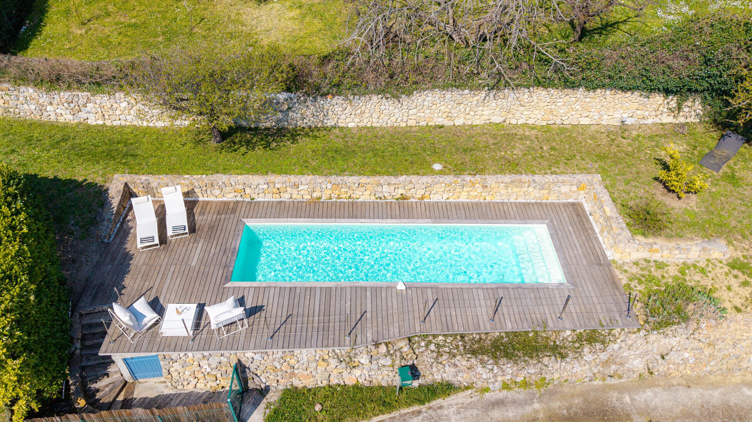 Una piscina rectangular con muelle de madera, rodeada por un muro de piedra y equipada con sillas de descanso y una pequeña mesa.