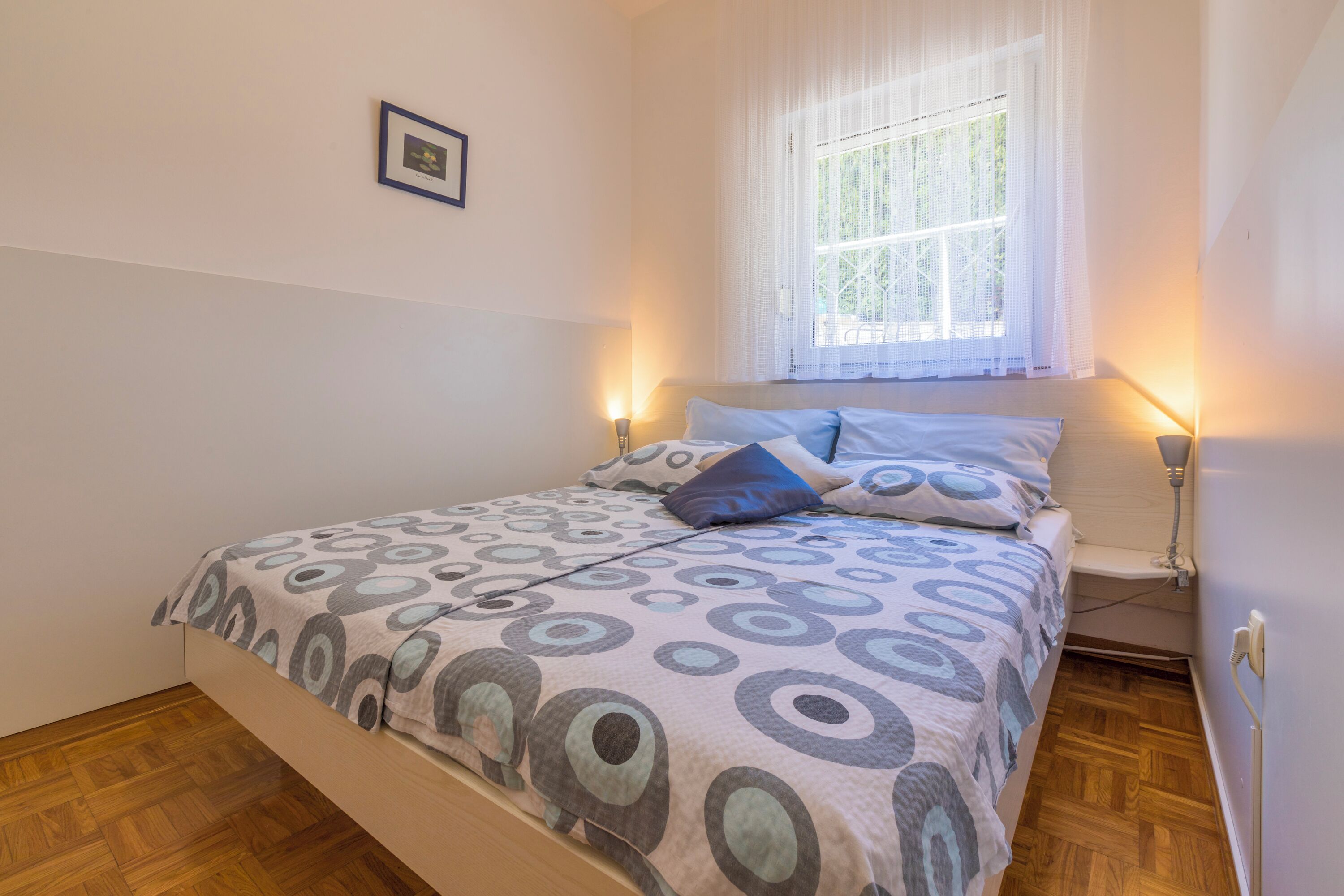 A bedroom with a bed, a window with curtains, a bedside table, and a wall-mounted picture.
