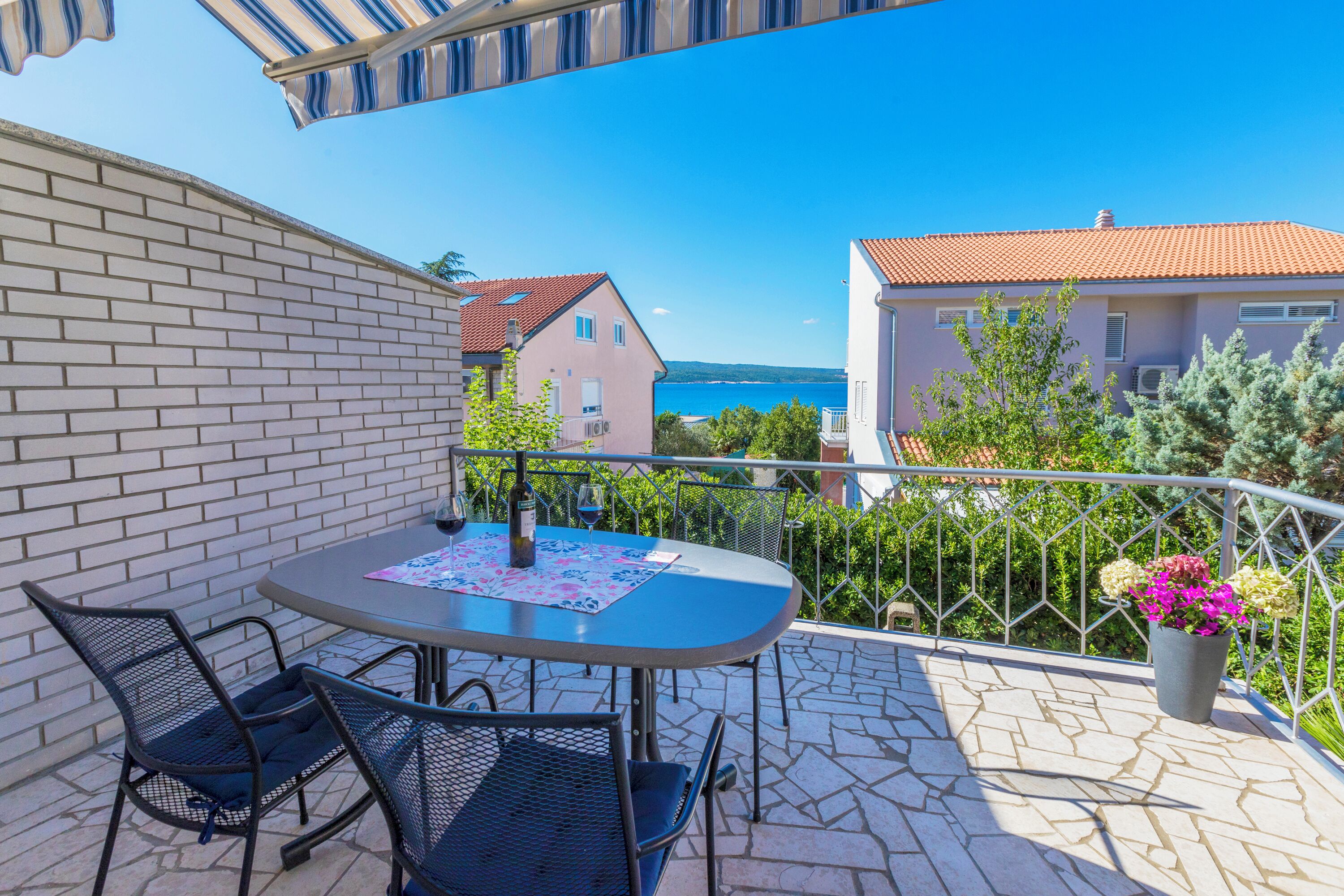 A balcony with a table and chairs, a potted plant, and a view of the sea and buildings.