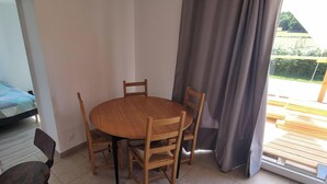 Quarto com uma cama, uma mesa redonda de madeira com cadeiras, uma janela com cortinas e vista para um deck de madeira lá fora.