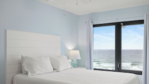 A bedroom with a bed, a nightstand, a lamp, and a view of the ocean through a window. - Island Sunrise 464 by Liquid Life (Gulf Shores)