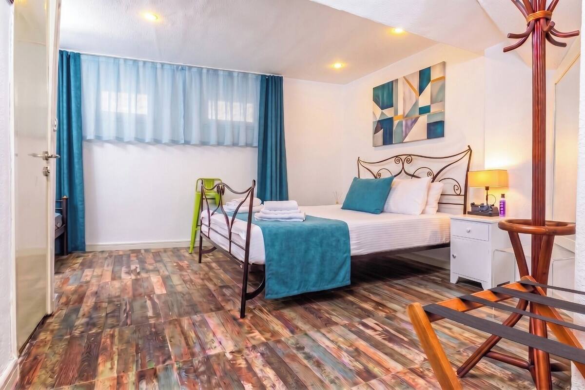 A hotel room with a bed, a bedside table, a lamp, a chair, a window with curtains, and a wooden floor.