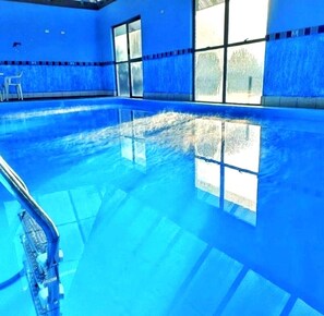 An indoor swimming pool with clear blue water, large windows, and a visible pool ladder.