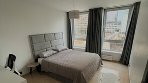 A bedroom with a bed, a desk, a chair, a small table, a rug, and a view of buildings through the window. - Paris La defense - Apartment 3 bedrooms (Courbevoie)