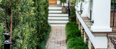A brick pathway flanked by neatly trimmed hedges leading to a white building with a balcony.