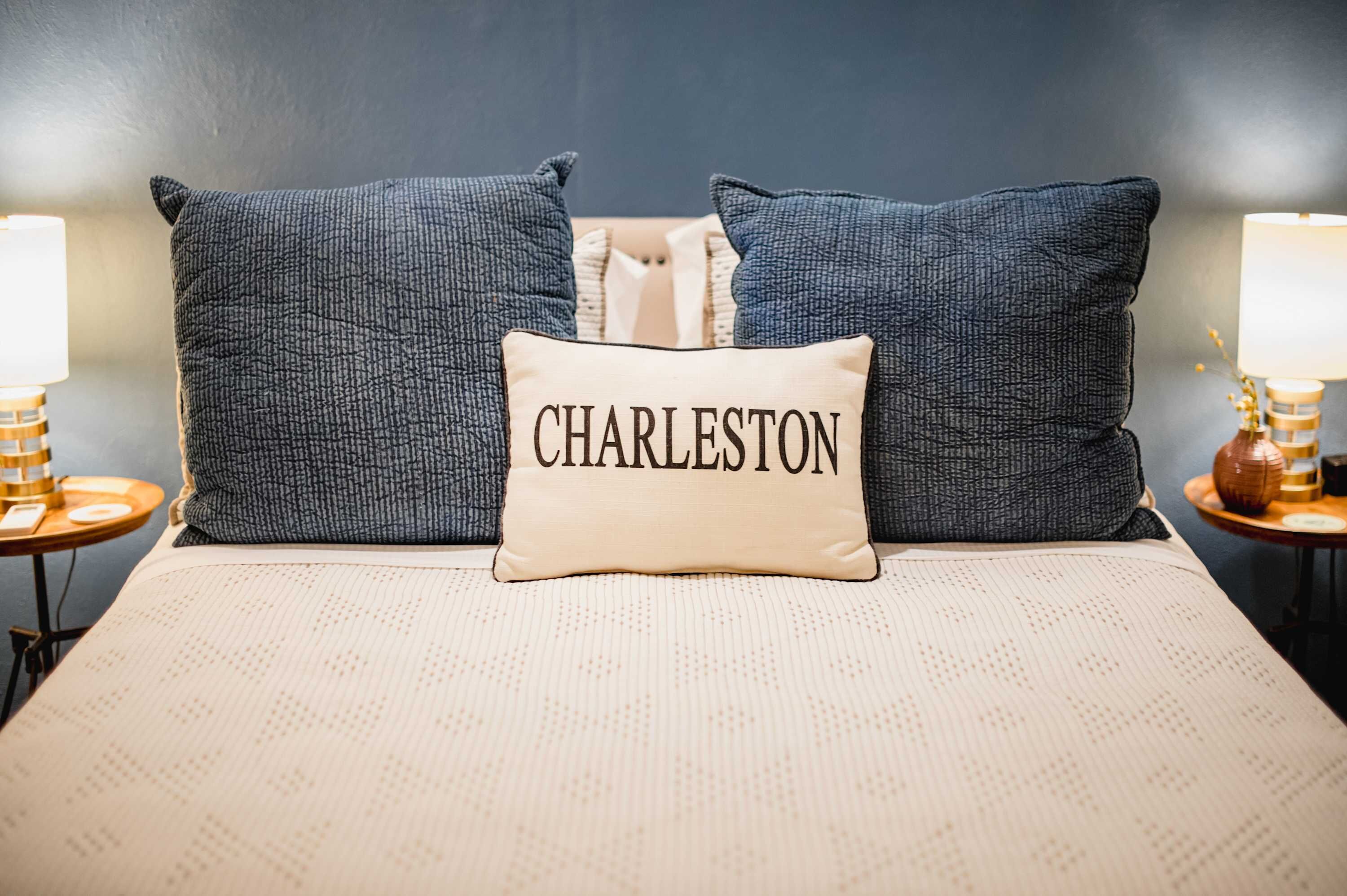 A bed with a pillow labeled 'CHARLESTON,' two matching pillows, a lamp, and a vase on a bedside table.