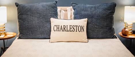 Un letto con un cuscino con l'etichetta 'CHARLESTON', due cuscini coordinati, una lampada e un vaso su un comodino.