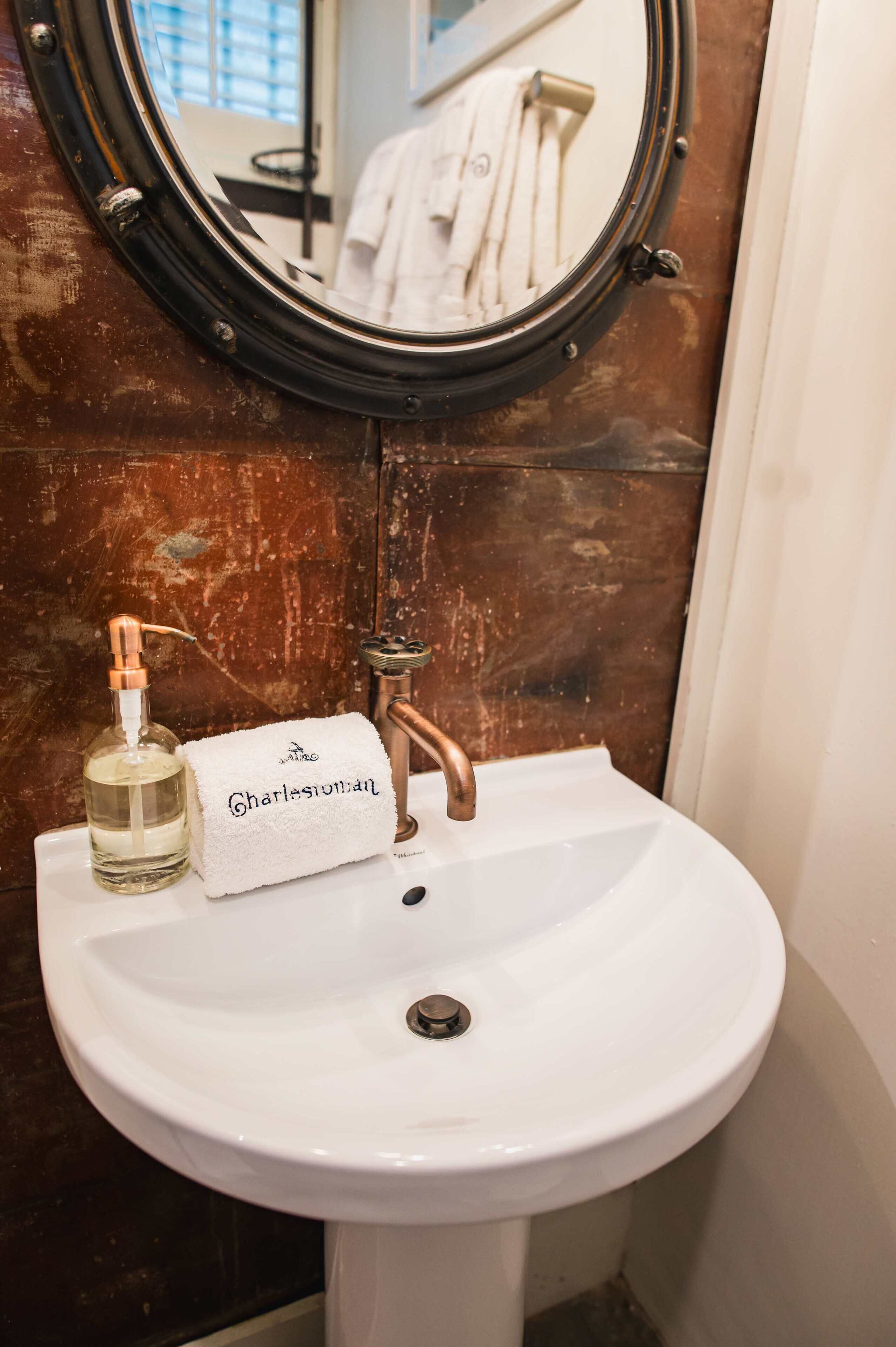 A white bathroom sink with a round mirror, a copper faucet, and a bottle of perfume.
