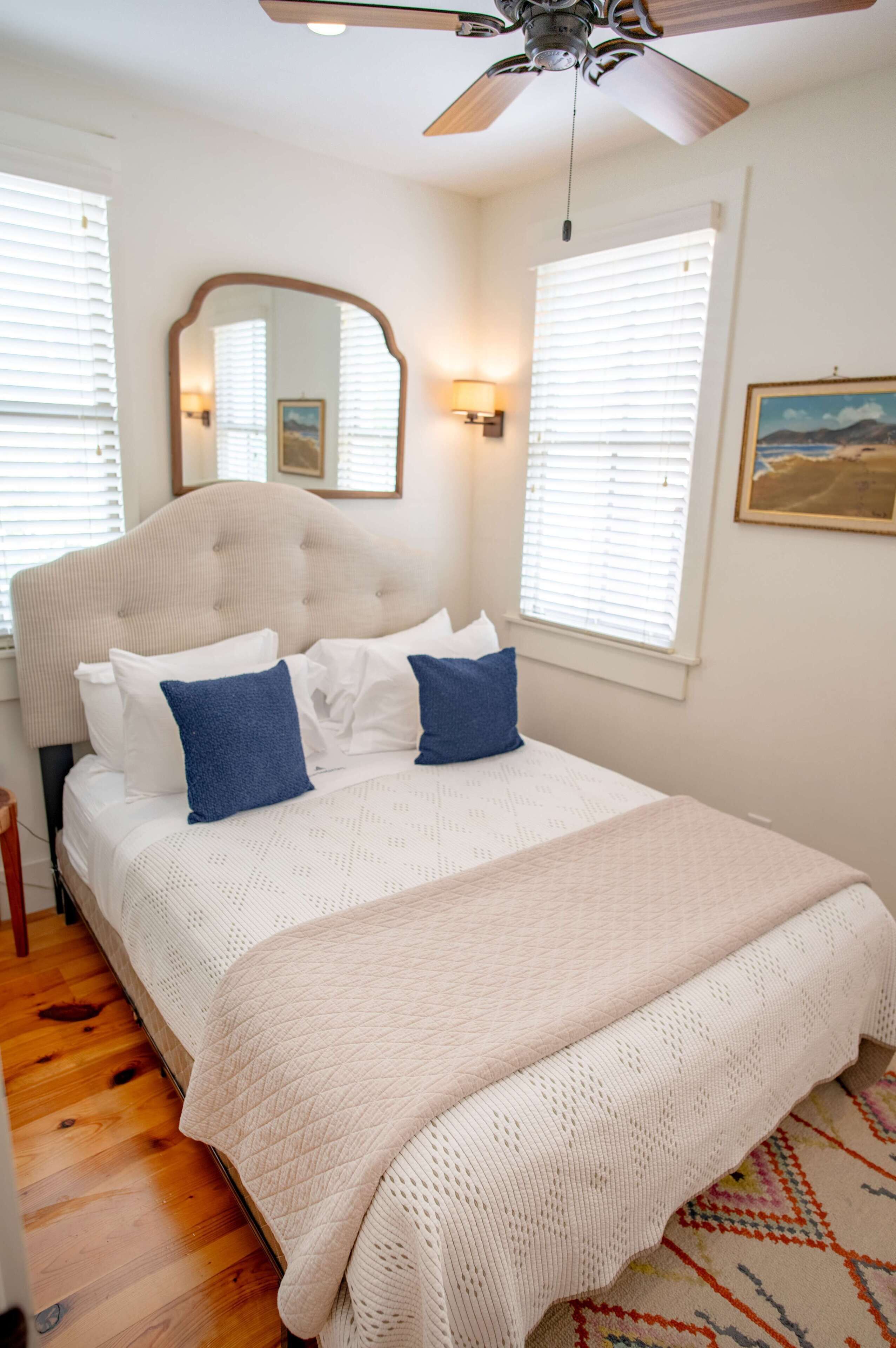A bedroom with a bed, a ceiling fan, a large mirror, and two windows with blinds.