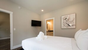 A bedroom with a bed, a flat-screen TV, a painting, and a bathroom visible through an open door. - Modern Upper Retreat w Free Tix Deck 2 Suites (Hendersonville)