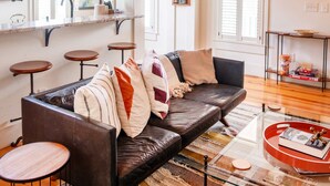 A spacious living area with a high wooden ceiling, a leather sofa, a glass coffee table, and a kitchen area with a window and shelves. - Walk Everywhere! Stylish 3BR Top Floor w/ Views (Charleston)