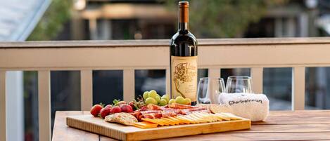Una mesa de madera con una botella de vino, copas y una tabla de embutidos en un balcón.