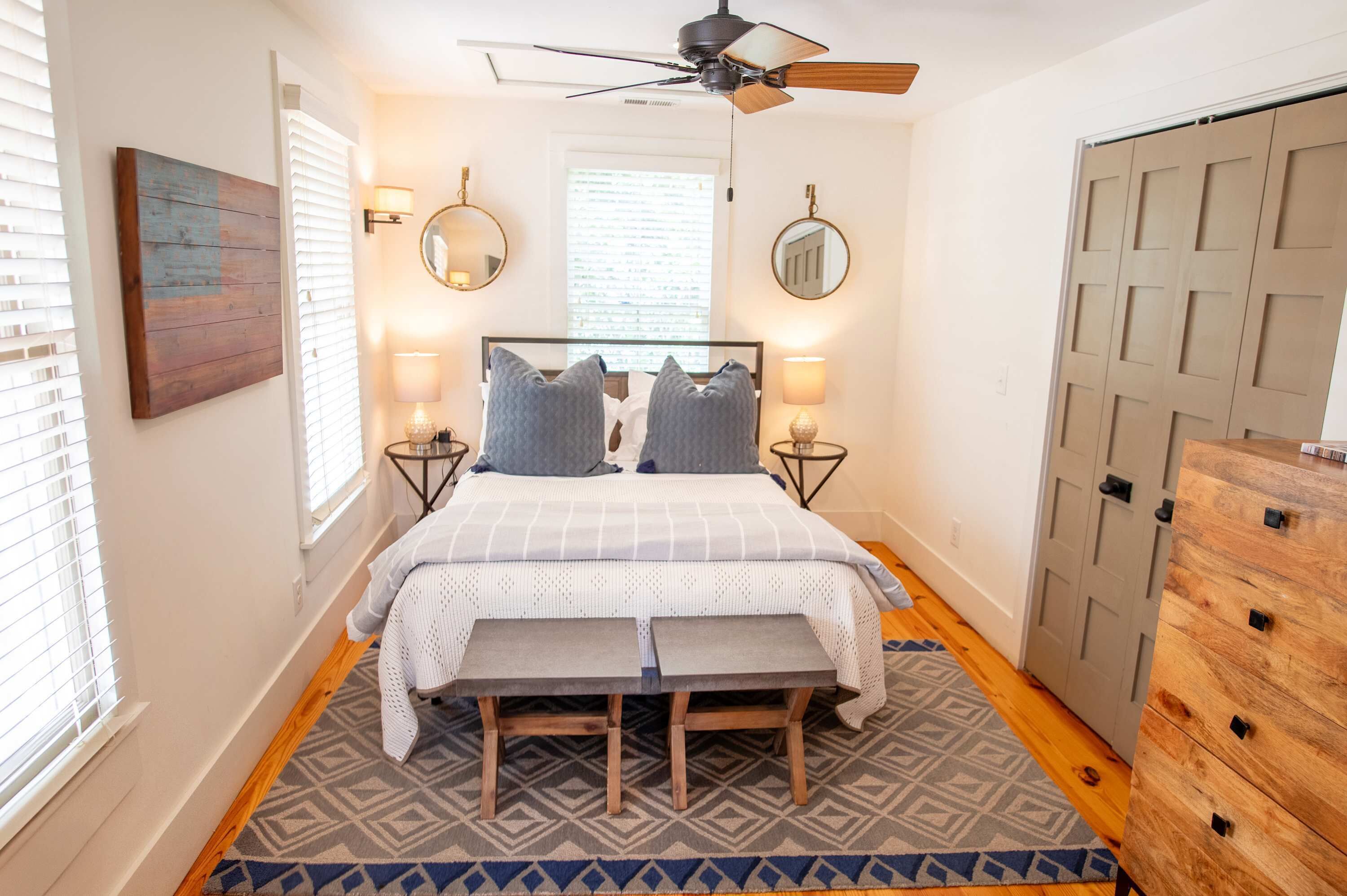 A bedroom with a bed, bedside lamps, a ceiling fan, a wooden dresser, and a patterned rug.