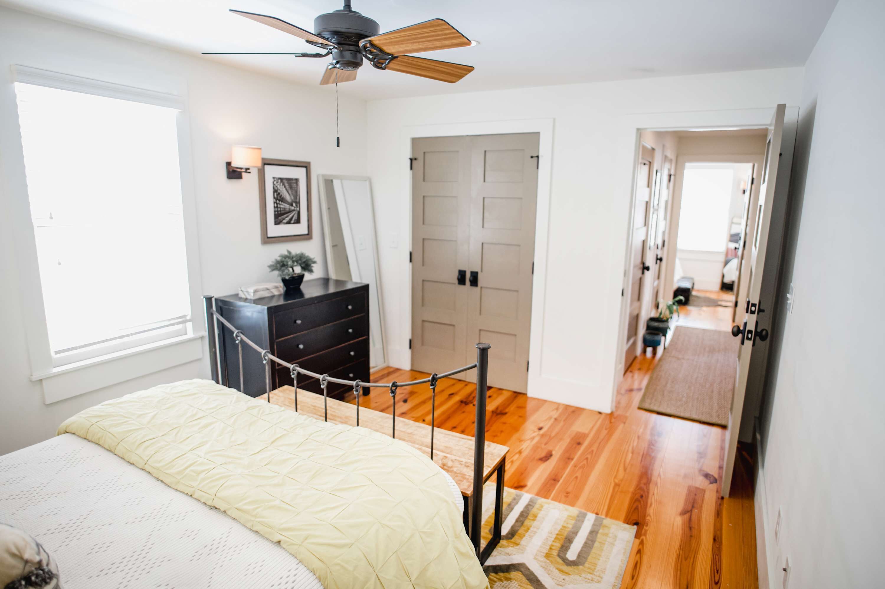 Une chambre à coucher comprenant un lit, une commode, un ventilateur de plafond, une porte et un couloir menant à une autre pièce.