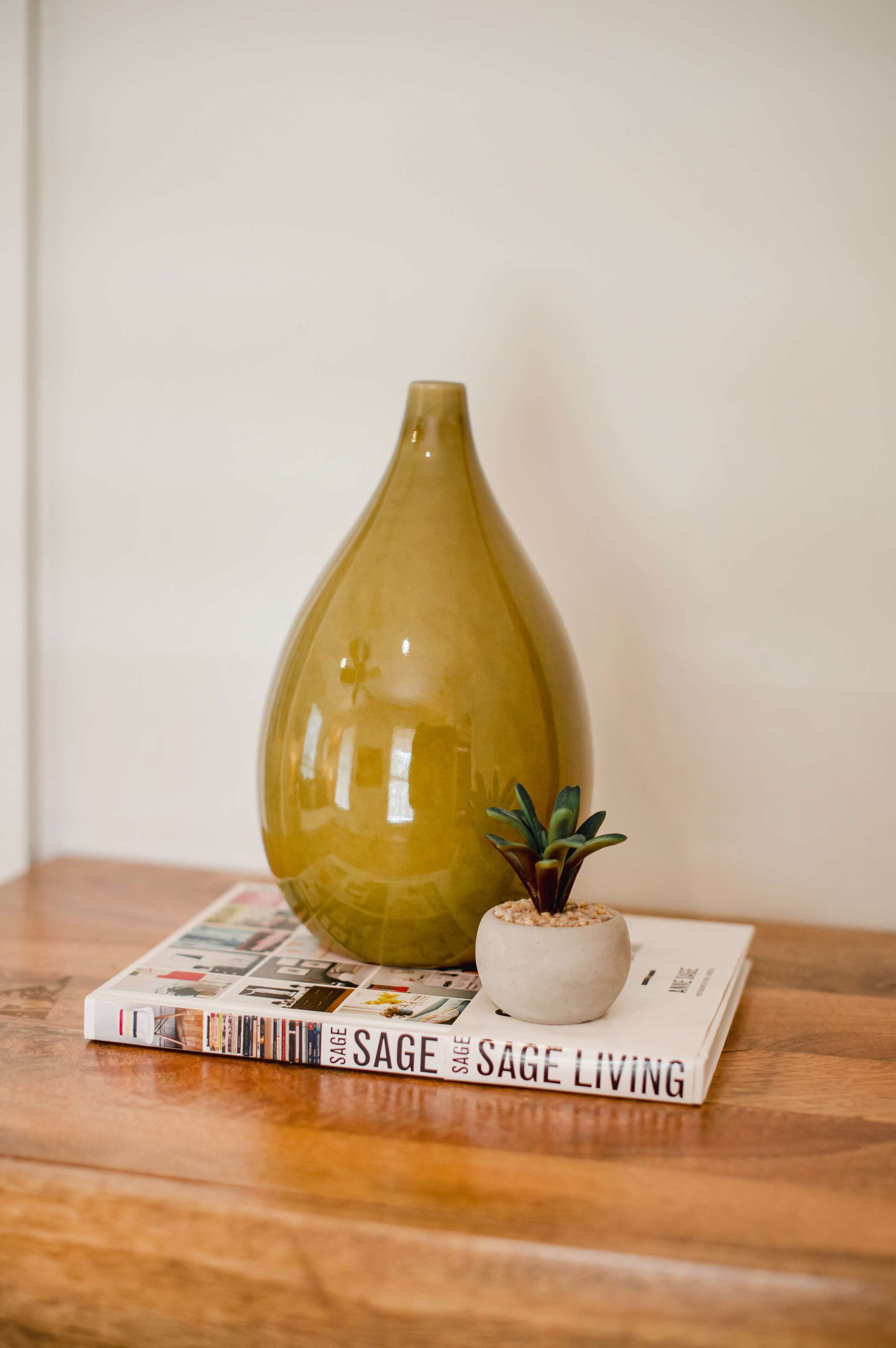 Un vase posé sur une surface en bois, à côté d’un livre intitulé « SAGE LIVING » et d’une petite plante.