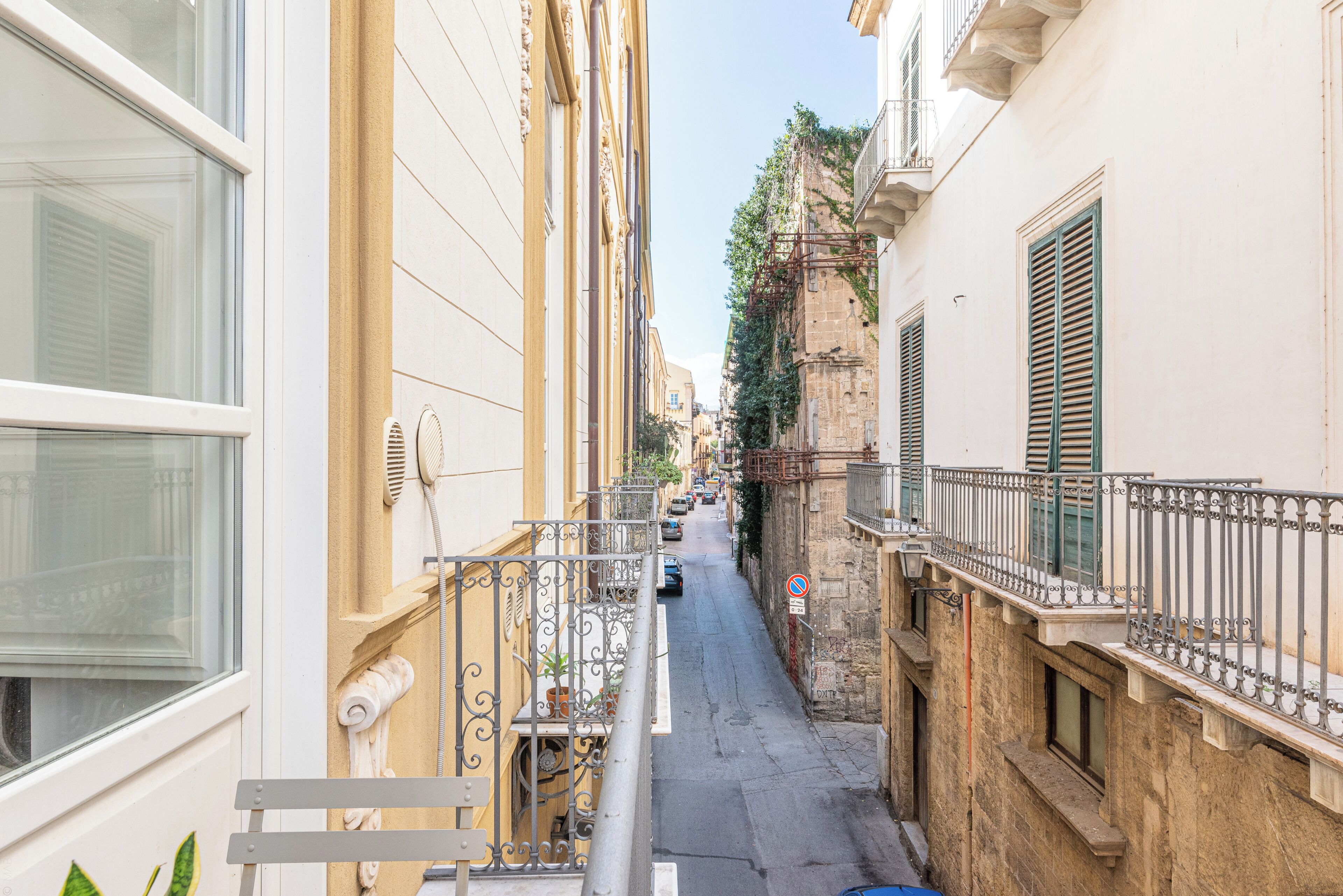 Une rue étroite, bordée de bâtiments des deux côtés, avec un balcon bien visible et un ciel dégagé.
