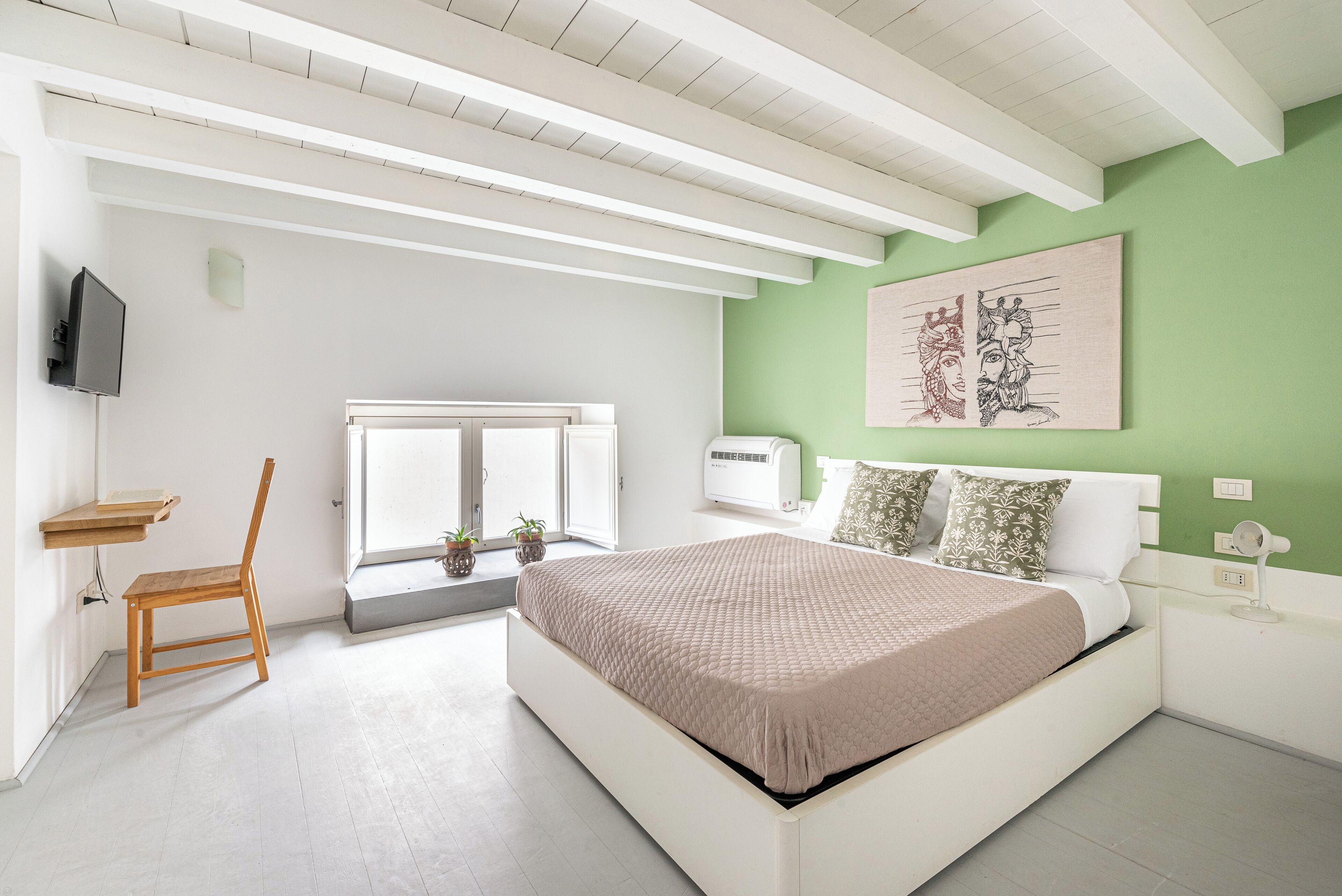 A bedroom with a bed, a chair, a window, and a wall-mounted TV.