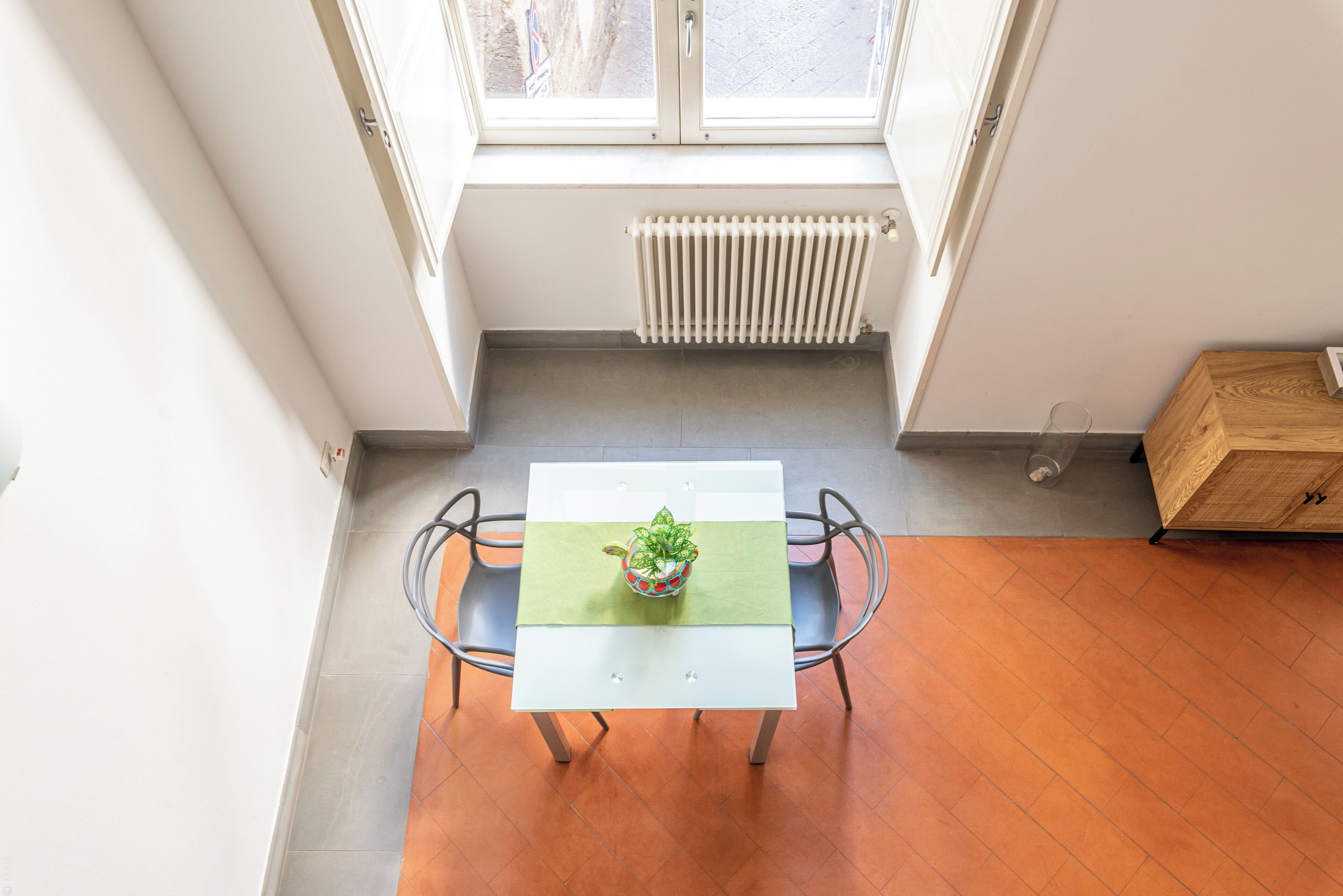 Une pièce avec une table blanche, deux chaises, une plante en pot, un radiateur et une fenêtre.