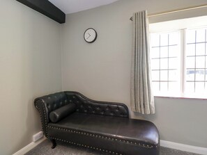 Una habitación con un sofá de cuero oscuro, un reloj de pared, una ventana con cortinas y un enchufe en el suelo.