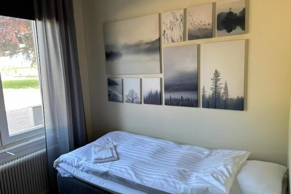 A single bed with a white mattress and a neatly folded towel, a window with curtains, and a wall with six landscape paintings.