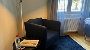 A room with a dark armchair, a small table with a water bottle and a magazine, a floor lamp, and a view of trees through a window. - Söderåsen Airport Hotel (Ljungbyhed)