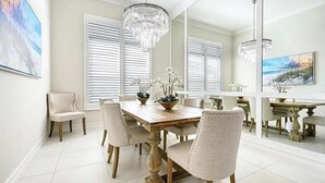 A dining room with a wooden table, white chairs, a chandelier, and large windows with blinds. - Luxury Naples Retreat / Pool, Near Beach (Naples)