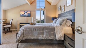A bedroom with a sloped ceiling, a bed with a patterned bedspread, a desk with a chair, and a fireplace. - 4073 Whale Watcher's Cove - Bay View Home (Monterey)