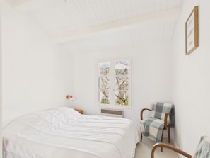 Ein Schlafzimmer mit Bett, Sessel, Fenster mit Blick auf Bäume und einem an der Wand hängenden Bilderrahmen.