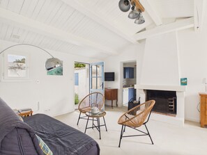 Ein geräumiges Wohnzimmer mit einer Couch, zwei Korbstühlen, einem Kamin und einem durchgehenden Gang sichtbarem Küchenbereich.