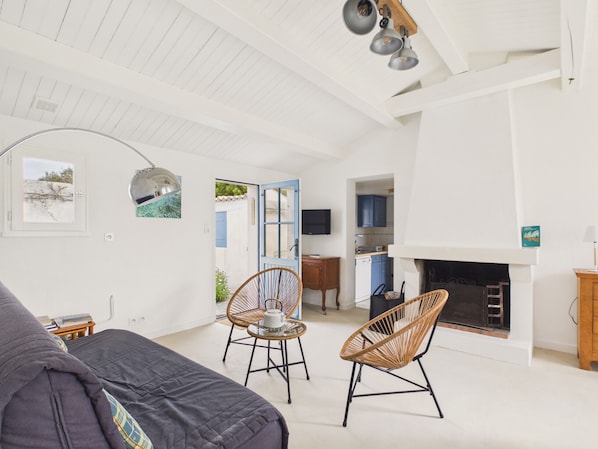 Ein geräumiges Wohnzimmer mit einer Couch, zwei Korbstühlen, einem Kamin und einem durchgehenden Gang sichtbarem Küchenbereich.