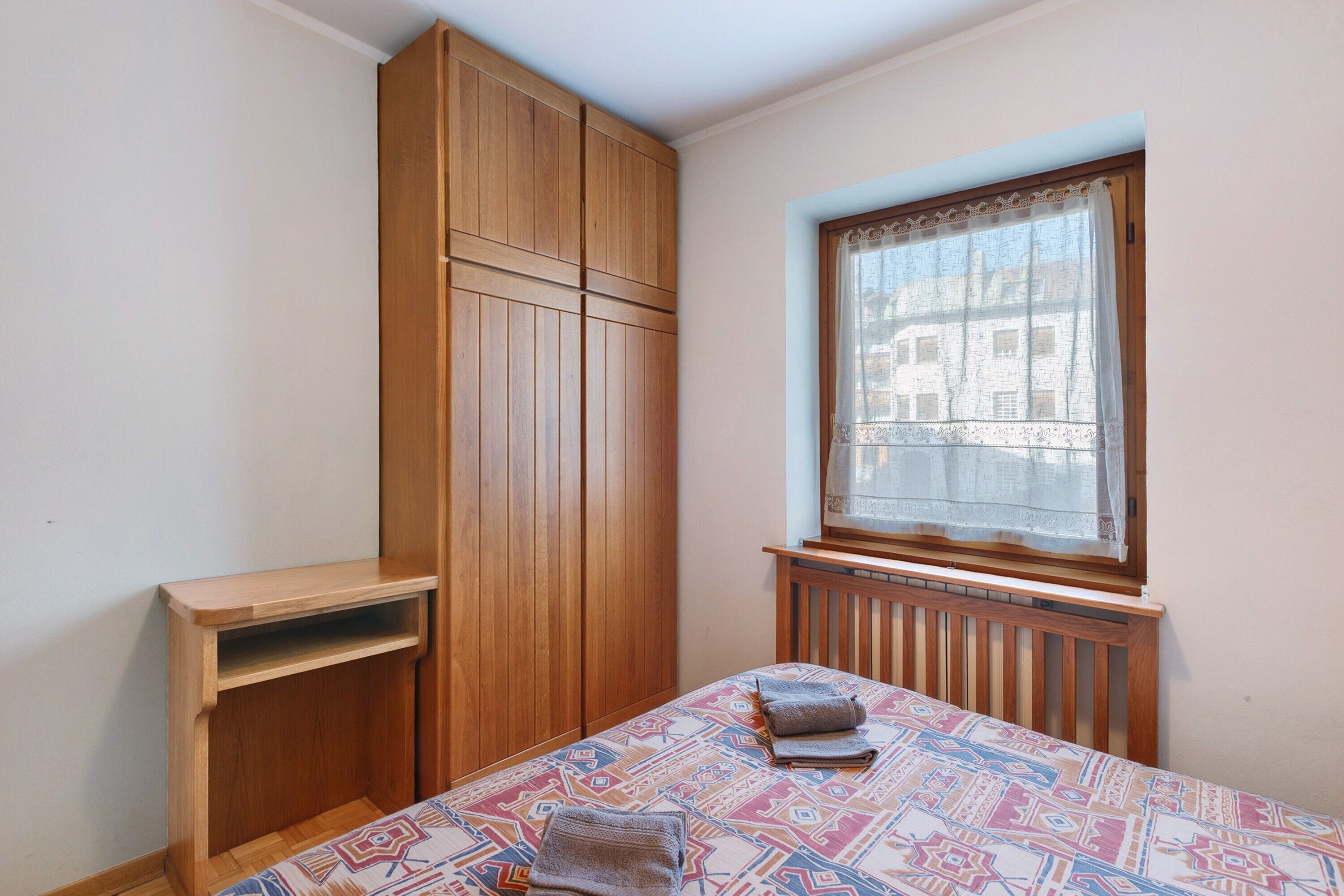 Ein Schlafzimmer mit einem Bett, einem Holzschrank, einem Fenster mit Spitzenvorhang und einer Heizung.