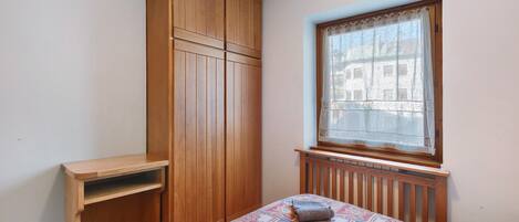 Ein Schlafzimmer mit einem Bett, einem Holzschrank, einem Fenster mit Spitzenvorhang und einer Heizung.