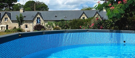 A clear blue swimming pool with a hexagonal tile pattern, surrounded by stone buildings with pitched roofs and a clear sky above.