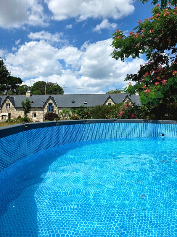 Une piscine d’un bleu éclatant, dont le fond est recouvert de carreaux hexagonaux, entourée de bâtiments en pierre aux toits en pente, sous un ciel dégagé.
