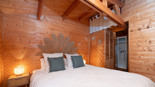 Een slaapkamer in een houten blokhut met een bed, kussens, een nachtkastje met een lamp en een houten plafond.