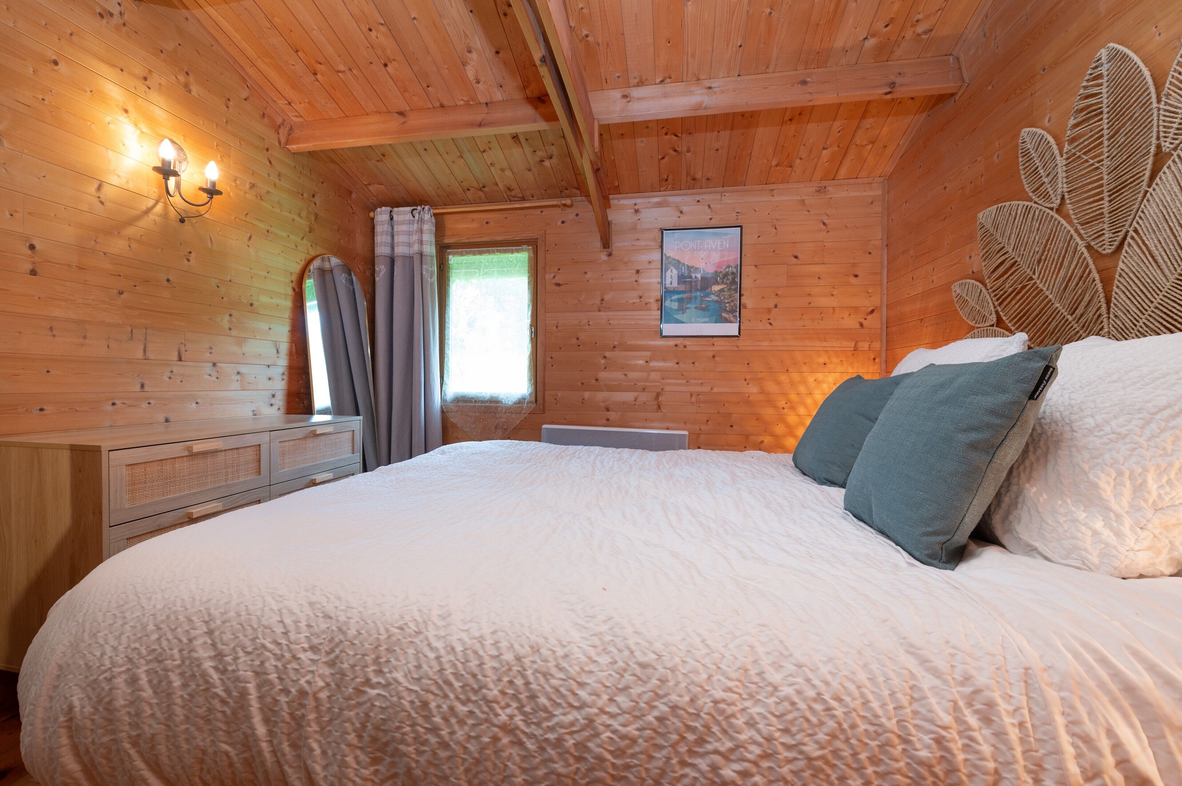 Une chambre dans une cabane en bois, avec un lit, une commode, une fenêtre avec des rideaux et un luminaire fixé au mur.