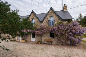 Una casa de piedra con camino de grava, un banco y enredaderas moradas en flor.