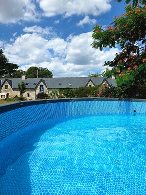 Une piscine d’un bleu éclatant, dont le fond est recouvert de carreaux hexagonaux, entourée de bâtiments en pierre aux toits en pente, sous un ciel dégagé.