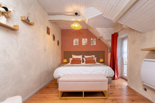 Un dormitorio con techo inclinado, una cama con cabecera, mesitas de noche, un banco y una ventana con cortinas.