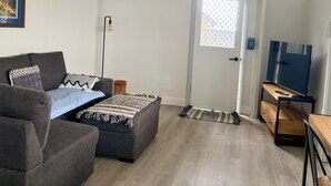 A living room with a sofa, ottoman, and a small rug. There is a door with a window, a wall-mounted air conditioner, and a lamp. - Takura Cottage On The Hill with Panoramic Views (Takura)