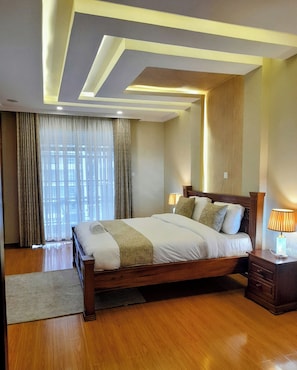 A hotel room with a wooden bed, a nightstand with a lamp, a large window with curtains, and a modern ceiling design. - Luxurious 4-bedroom Apartment With Pool Access in Lavington (Nairobi)