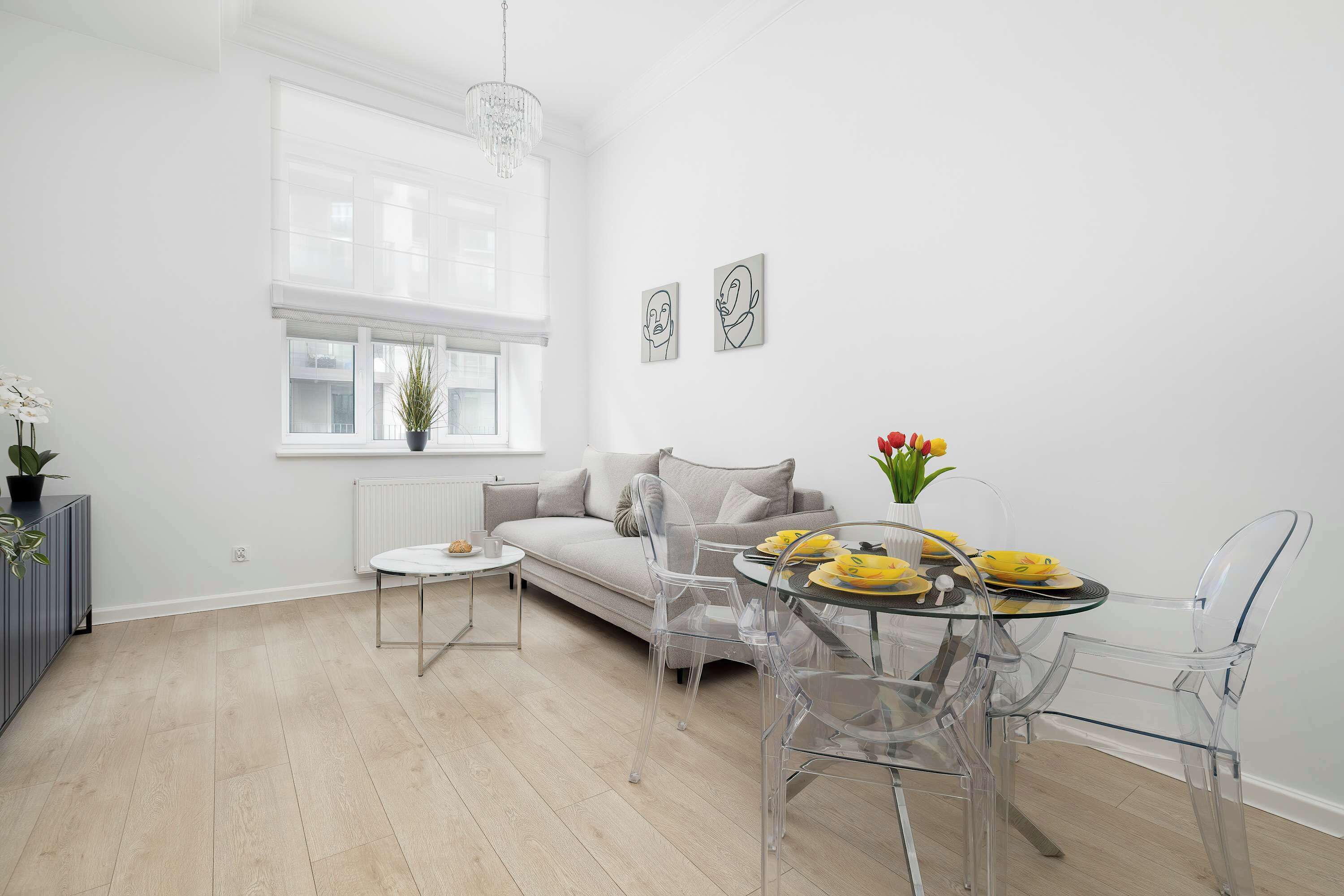 Una sala de estar moderna con un sofá, sillas traslúcidas, una mesa de comedor con platos amarillos y una ventana con persianas.