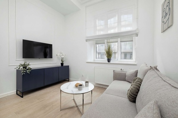 A modern and minimal living room setup with a grey fabric sofa, a dark blue media console with a TV on top, and two round coffee tables. The space is bright and airy with large windows.