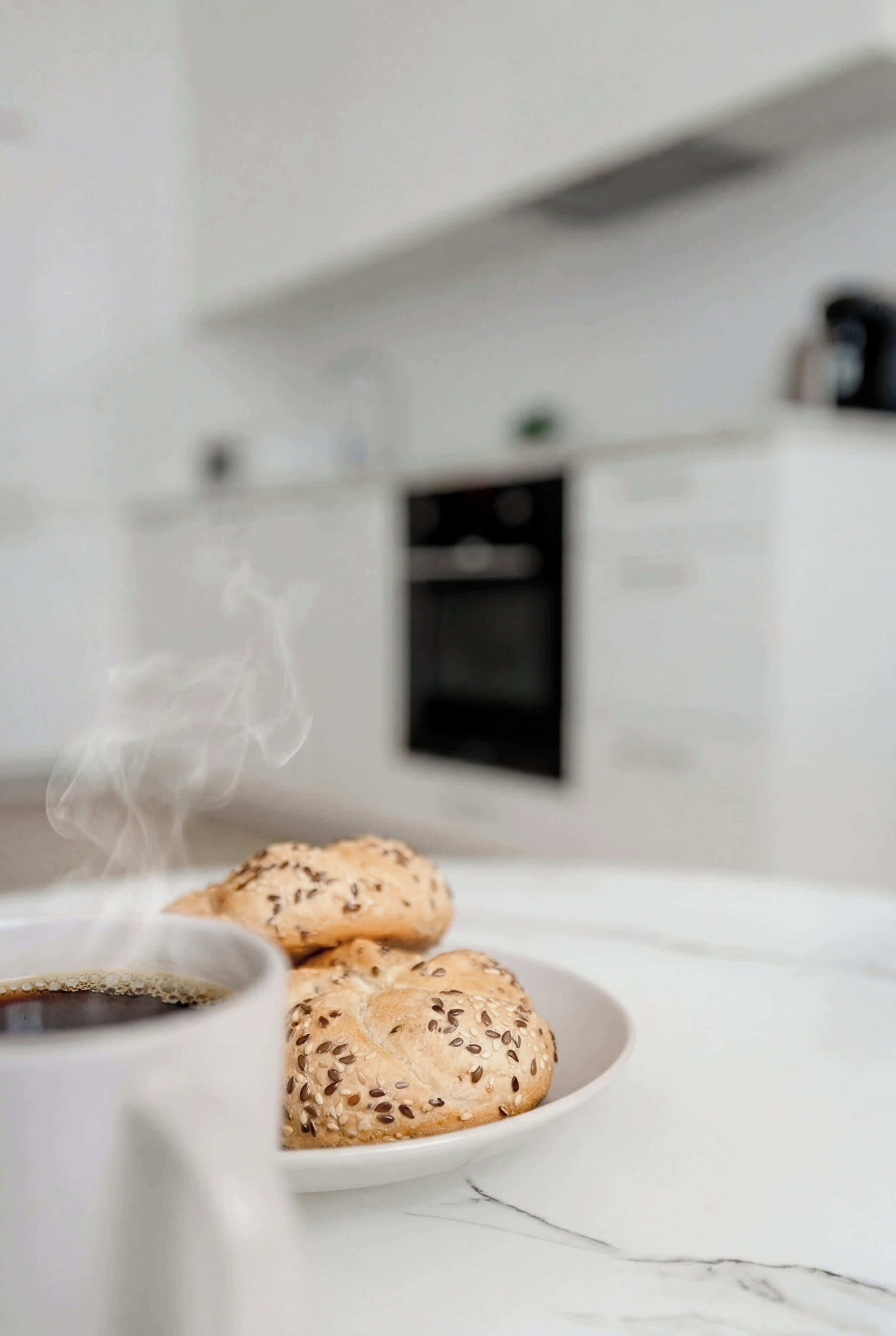 A white ceramic mug with a beverage, a plate with sesame seed buns, and a kitchen with white cabinets and a black oven in the background.