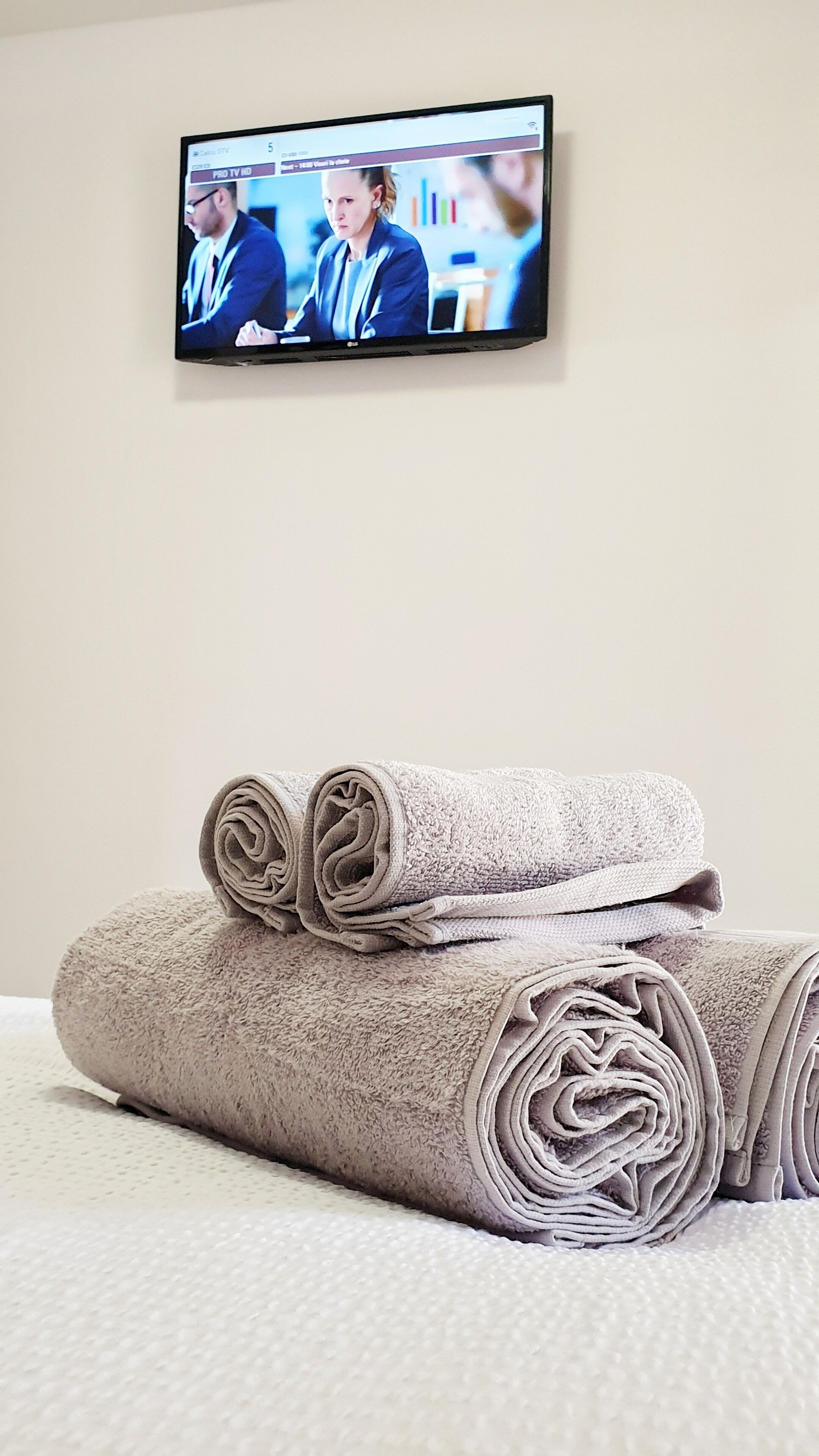 A television mounted on a wall displaying a news program, with two rolled towels placed on a bed.