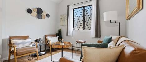 Una sala de estar moderna con un sofá de cuero, una silla de madera, una mesita de centro y un ventanal con cortinas.