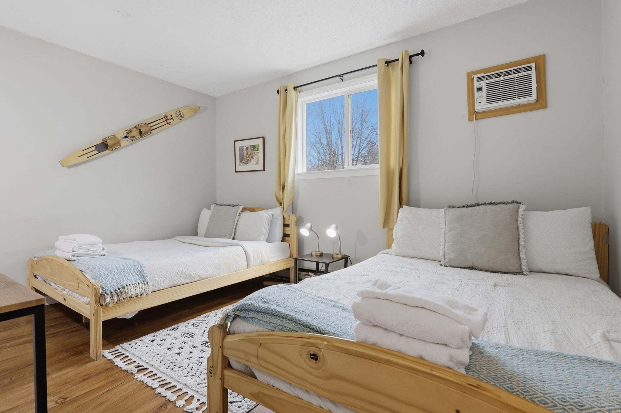 Un dormitorio con dos camas, piso de madera, ventana con cortinas, mesita de noche con lámpara y aire acondicionado instalado en la pared.
